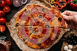 Eating pizza, top view. Hand taking slice of hot delicious pizza, selective focus. Pizza ingredients on the wooden table