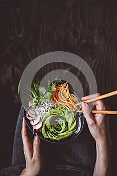 Eating raw vegan bowl with rice noodles, vegetables and avocado on wooden background. Top view with copy space