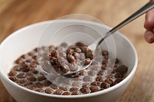 Eating with spoon chocolate cereal balls with milk in white bowl for breakfast on wooden table