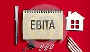 EBITA text writtenon red background on a notebook with pencils and office tools and model wooden house, business