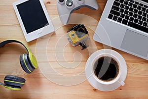 Ebook reader and wireless headphones and other electronic gadgets on wooden desk with laptop computer with black coffee in white