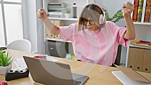 Ebullient young hispanic woman, glowing with success, celebrating a business win while working on a laptop, listening to music on