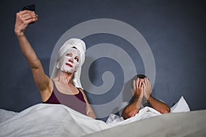 Eccentric housewife with makeup facial mask and towel taking selfie in bed and husband with desperate face expression in weird man