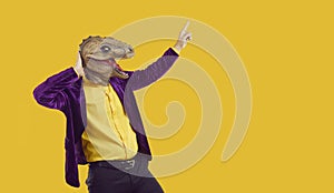 Eccentric showman in dinosaur mask and velvet jacket pointing index finger on yellow background.