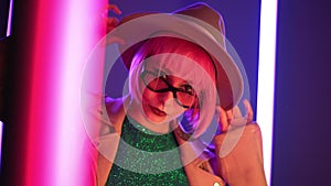 Eccentric woman with pink hair posing near led-colorful neon lamps. Charming odd girl in hat and sunglasses, nightlife