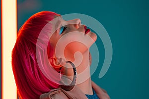 Eccentric woman with pink hair posing near led neon lamp on teal background. Charming odd girl, nightlife concept