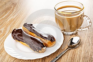 Eclairs with chocolate glaze in plate, cup of coffee with milk, spoon on wooden table
