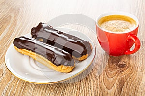 Eclairs with chocolate glaze in plate, cup with coffee on wooden plate