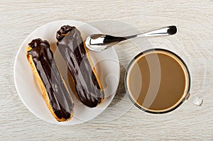 Eclairs with chocolate glaze in plate, spoon, cup of coffee with milk on wooden table. Top view