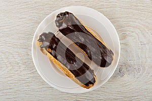 Eclairs with chocolate glaze in white plate on wooden table. Top view