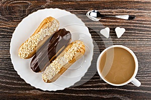 Eclairs with chocolate and peanut in plate, spoon, pieces of sugar, coffee with milk in cup on wooden table. Top view