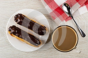 Eclairs with chocolate in white plate, spoon on napkin, cup of coffee with milk on wooden table. Top view