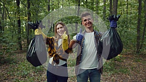 Eco activism concept. Young man and woman volunteers showing garbage bags and smiling to camera, gesturing thumbs up