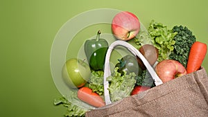 Eco bag with assorted raw organic vegetables and fruits on green background.