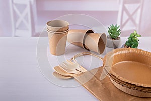 Eco friendly craft paper dishware and wooden forks, spoons in kitchen. Cups, plates, fork, spoon, bag on white table.