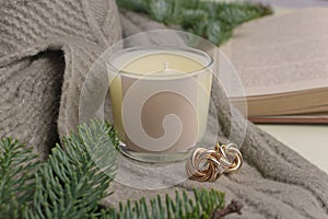 Eco-friendly handmade soy wax candle in a glass candlestick. Nearby lies a decor of fir branches, a warm sweater, a book and