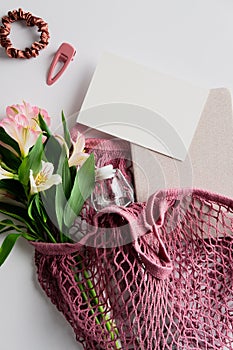 Eco-friendly string shopping bag with fresh flowers, glass bottle, white paper card mockup, notebook, feminine accessories