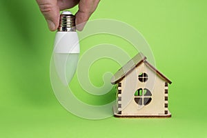 Eco power concept. LED bulb in hand and toy wooden house on green background