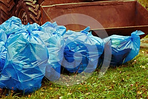 Pile of blue trash bags filled with garbage on the grass and the tractor bucket