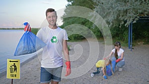 Ecology rescue, portrait of smiling activist man in rubber gloves with garbage bag near pointer sign on background wife