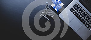 Ecommerce. Laptop computer, shopping trolley and white gift with blue ribbon on dark background. Website retail business