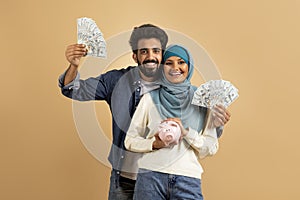 Economy Concept. Happy Young Muslim Couple Holding Cash And Piggy Bank