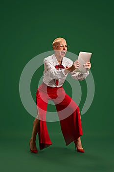 Ecstatic businesswoman dressed in fashion white shirt, red pants, holding tablet against green studio background.