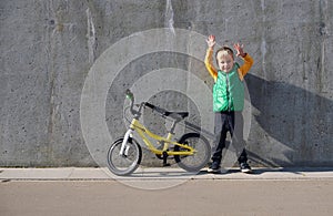 Ecstatic funny little child boy standing near concrete wall with his yelllow bicycle on sunny day. Urban lifestyle