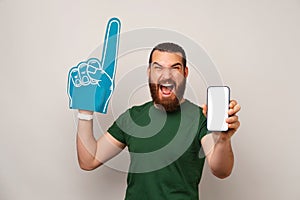 Ecstatic man wearing foam finger fan glove while showing blank phone screen.