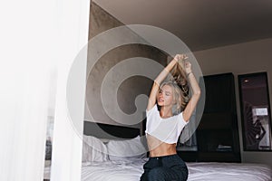 Ecstatic slim girl stretching in morning. Blithesome blonde lady sitting on bed with hands up