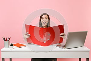 Ecstatic woman looking on red folder with paper documents, working on project while sitting at office with laptop