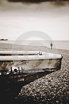 Edge of old wooden boat with lonely female figure on pebble beach and remains of the west pier in distance and overcast sky, Brig