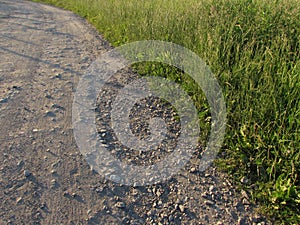 The edge of the roadside of a light rocky forest road and green summer grass in a corner