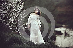 On the edge of the shore near the river there is a girl in a white dress near a flowering tree. The girl in the wind in