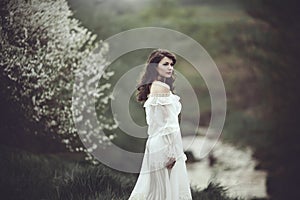 On the edge of the shore near the river there is a girl in a white dress near a flowering tree. The girl in the wind in