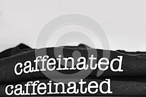 The edge of two folded black tee-shirts with the word caffeinated from both showing folded and stacked at bottom of frame with photo