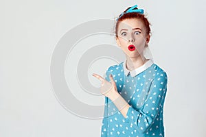 Edhead girl in blue dress, pointing at blank studio wall, surprised with sale prices, keeping her mouth wide open.