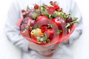 Edible bouquet consisting of pomegranate, apples, plums and scarlet roses in hands of woman on white background