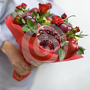 Edible bouquet consisting of pomegranate, apples, plums and scarlet roses in hands of woman on white background