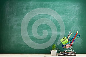 Education concept - school supplies in a shopping cart on the desk in the auditorium, blackboard background