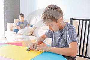 Education, online learning, entertiment at home. Boy making paper craft card with colored paper. Brother using tablet photo