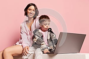 Educational games. Young woman and little boy, mom and son using laptop isolated on pink studio background. Mother's