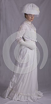 Edwardian woman in white blouse, skirt and hat standing against a studio backdrop