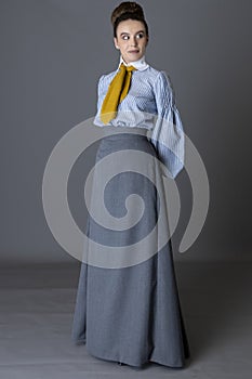 An Edwardian working class woman wearing a striped cotton blouse, mustard yellow cravat, and a grey walking skirt