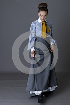 An Edwardian working class woman wearing a striped cotton blouse, mustard yellow cravat, and a grey walking skirt