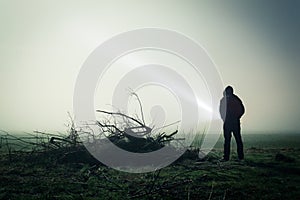 An eerie silhouette of a lone hooded figure in a field on a foggy night with a torch. With a dark edit