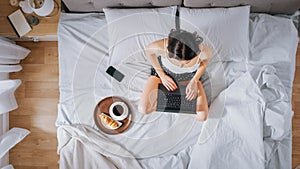 Efficient Young Millennial Girl Sitting on a Bed in the Morning, Uses Laptop Computer and Eats Cro
