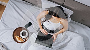 Efficient Young Millennial Girl Sitting on a Bed in the Morning, Uses Laptop Computer and Eats Cro