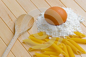 Egg in flour with penne and wooden spoon on wooden background