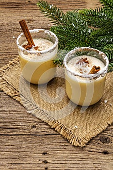 Eggnog cocktail in two glasses arranged with christmas decoration on old wooden table. Evergreen fer tree branch, artificial snow